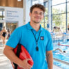 junger sportlicher Mann steht mit einer Rettungsboje unterm Arm am Beckenrand eines Schwimmbads